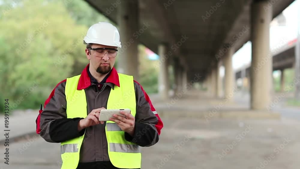Workman in hard hat uses smartphone under overpass. Manager of the repair works at construction site under overhead road inputs data in his smartphone Workman in hard hat uses smartphone under overpass. Manager of the repair works at construction site under overhead road inputs data in his smartphone