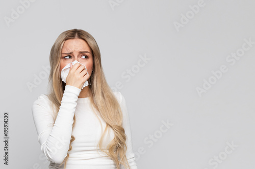 Studio portrait of cute unhealthy blonde female in white top with napkin blowing nose, looking aside/Sick desperate female has flu/Rhinitis, cold, sickness, allergy concept/Allergy to dust, pollen photo