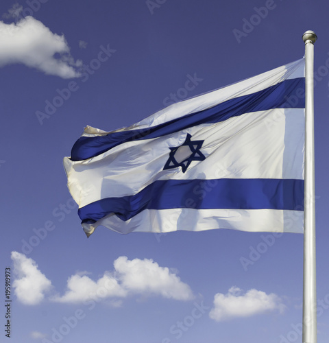 Israel flag flapping in the wind isolated against the blue sky. The flag is on a pole and flapping to the left. there are white clouds in the sky