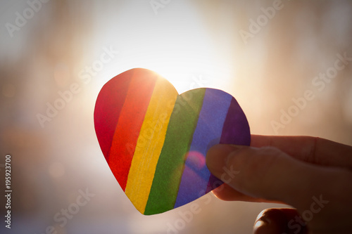 LGBT day concept, hand holds a heart painted like a LGBT flag, silhouetted against sun photo