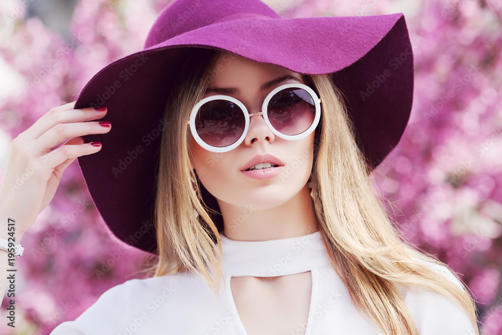 Outdoor close up portrait of young beautiful girl wearing stylish round  sunglasses, trendy violet hat, white shirt, posing in street. Pink blooming  trees on background. Spring fashion concept. Stock Photo | Adobe