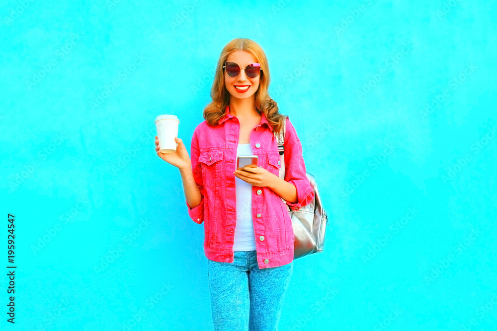 Fashion woman is using smartphone in pink denim jacket holds coffee cup on blue background Fashion woman is using smartphone in pink denim jacket holds coffee cup on blue background