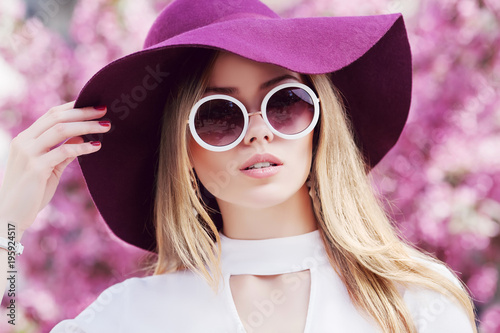 Outdoor close up portrait of young beautiful girl wearing stylish round sunglasses, trendy violet hat, white shirt, posing in street. Pink blooming trees on background. Spring fashion concept. 