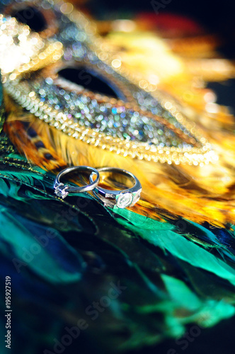 Wedding rings, jewelry and symbols attributes taken with selective focus on peacock colored carnival mask. Holiday, celebration. Macro. Blur. Bridal bouquet on background