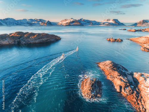 Fotografi Aerial view of fishing boats, rocks in the blue sea, snowy mountains and colorful sky with clouds at sunset in winter in Lofoten islands, Norway, Landscape with two ship, blue water, waves