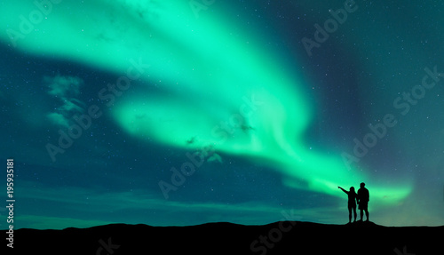 Fotografía Aurora borealis and silhouette of standing man and woman who pointing finger on northern lights