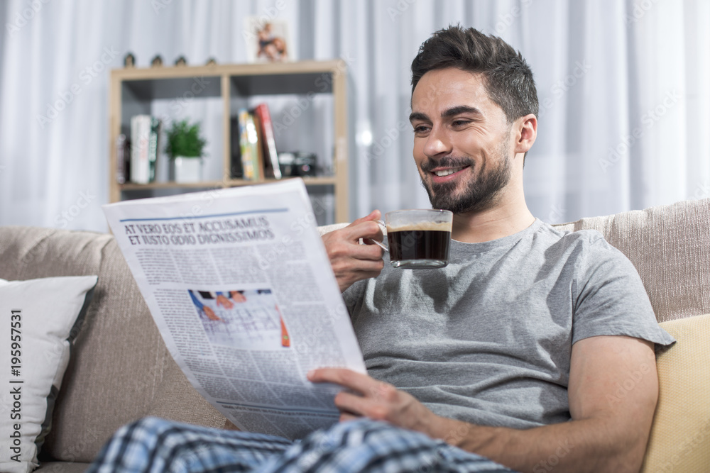 Φωτογραφία Portrait of contented handsome male reading news on couch and holding cup of cof