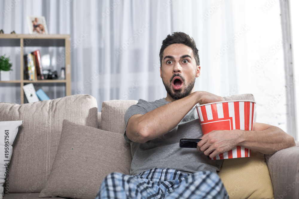 Φωτογραφία Waist up portrait of astonished guy sitting on sofa holding bucket with popcorn