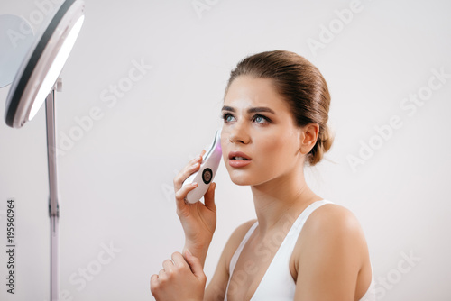 Attractive young woman with healthy porcelain skin and natural make-up is using the electric face massager in the front of cosmetic lamp at white background.