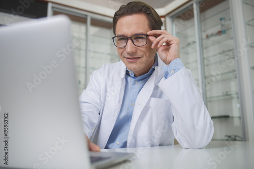 Portrait of smiling doctor typing in notebook computer while sitting at table indoor. Specialist during job concept