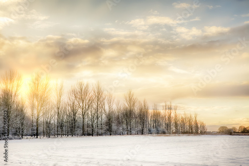 Cold and snow covered landscape on a winters morning with sunrise setting the sky on fire. Bare trees line the horizon and snow fields