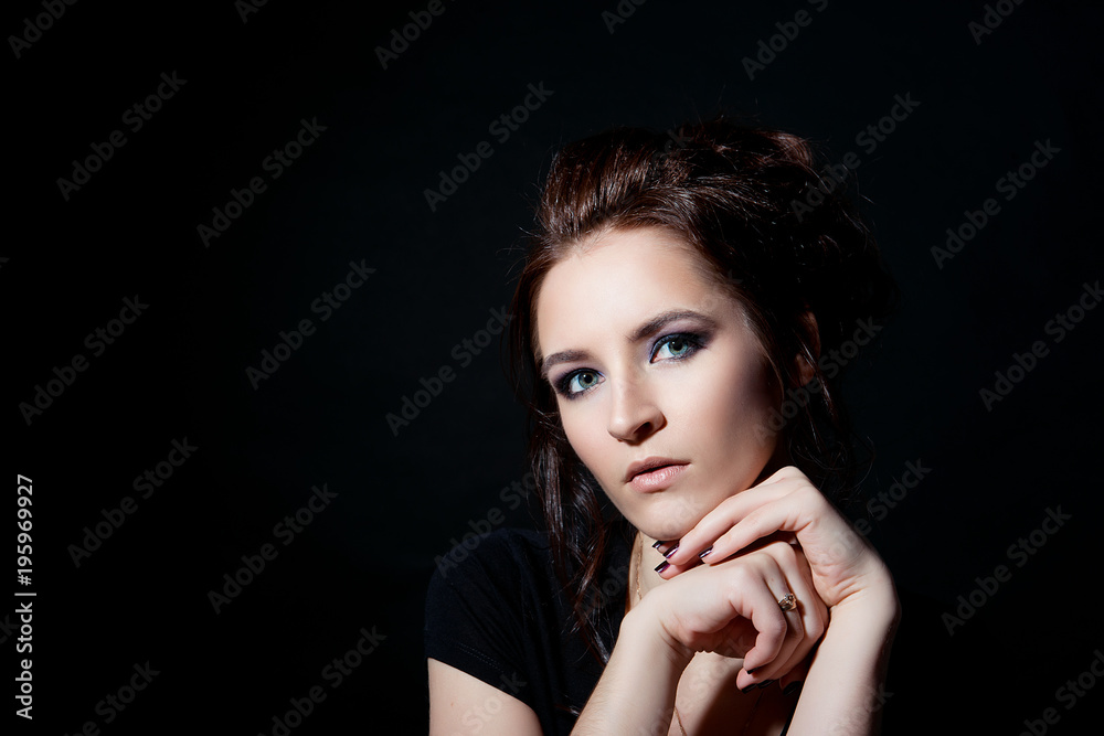 A brunette girl with evening make-up is holding hands under her chin close-up on a black background A brunette girl with evening make-up is holding hands under her chin close-up on a black background