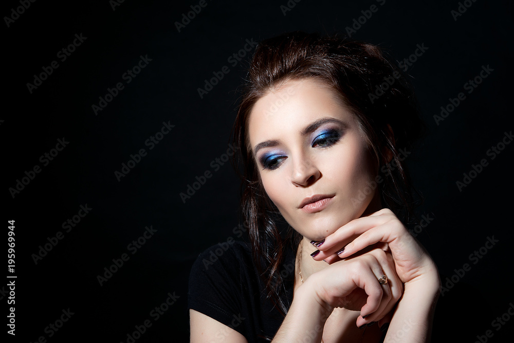 A brunette girl with an evening make-up is holding hands under her chin and is looking down a close-up on a black background A brunette girl with an evening make-up is holding hands under her chin and is looking down a close-up on a black background