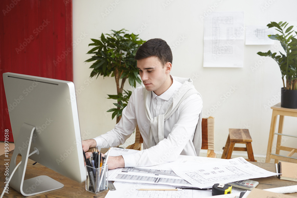Picture of concentrated male architect sitting at desk with project documentation and drawings, working in CAD system using generic computer. People, job, occupation, career and technology concept