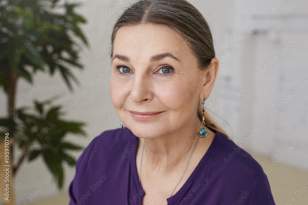 Portrait of serene mature woman with gray gathered hair, beautiful features and wrinkled skin posing indoors, looking at camera with happy smile, white wall and decorative plant in background Portrait of serene mature woman with gray gathered hair, beautiful features and wrinkled skin posing indoors, looking at camera with happy smile, white wall and decorative plant in background