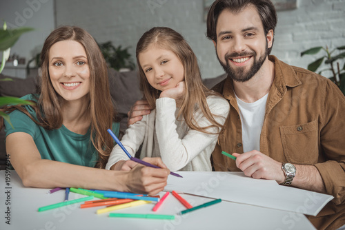 smiling parents and daughter drawing with felt-tip pens and looking at camera