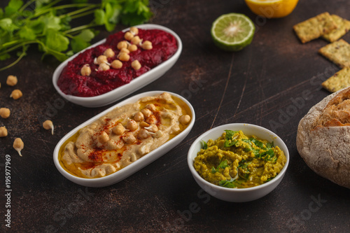 Vegan food background. Vegetarian snacks: hummus, beetroot hummus, green peas dip, vegetables. Top view, dark background, copy space.