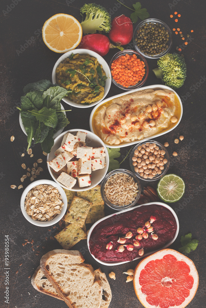 Vegan food background. Vegetarian snacks: hummus, beetroot hummus, green peas dip, vegetables, cereals, tofu. Top view, dark background, copy space.