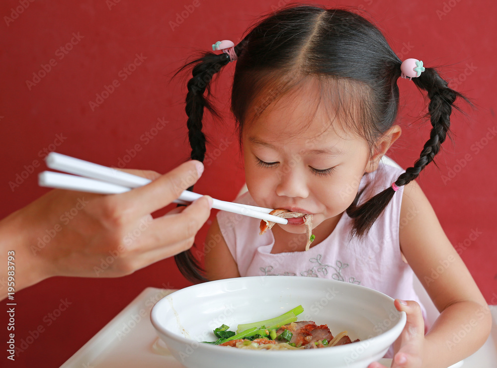 Mother feeding food for her daughter on high chair against red wall background. Mother feeding food for her daughter on high chair against red wall background.