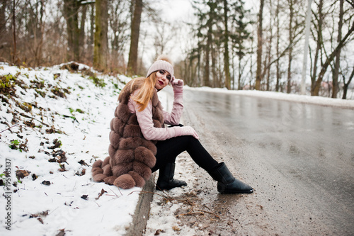 Stylish girl in fur coat and headwear at winter day on sitting border ofroad. photo