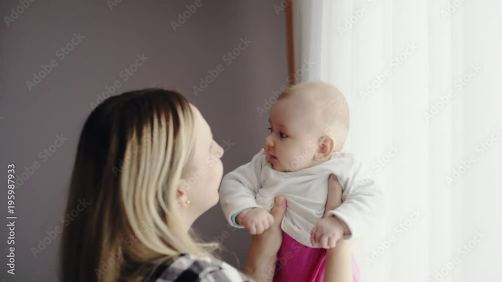 Happy loving mother playing with her young baby girl in a pink jumper suit lifting her in the air before lowering her to her chest for a hug Happy loving mother playing with her young baby girl in a pink jumper suit lifting her in the air before lowering her to her chest for a hug