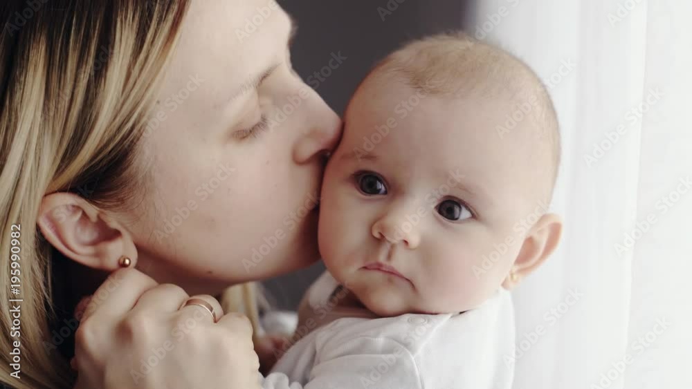 Loving mother kissing and nuzzling a young baby she is holding in her arms before turning to the side with a smile and laughing Loving mother kissing and nuzzling a young baby she is holding in her arms before turning to the side with a smile and laughing