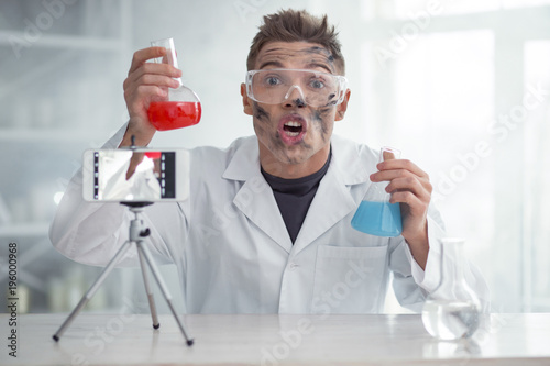 It is amazing. Handsome impressed dirty fair-haired young blogger and wearing uniform and holding test tubes while making new video