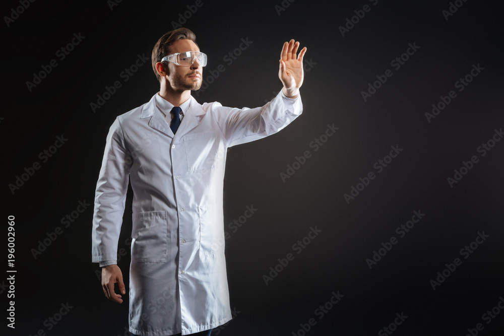 Personal task. Skilled smart handsome scientist standing in the dark room holding hand up and working.