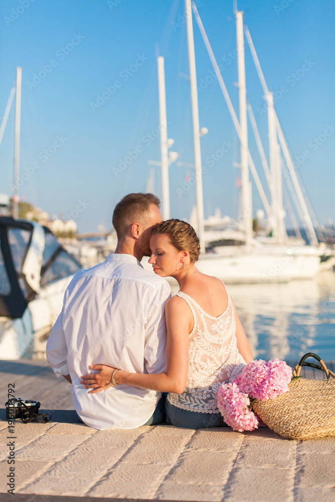 Couple of travelers are sitting in sea yacht resort during romantic honeymoon vacation at sunset. Man and woman are tourists. Lovers have pink hydrangea flowers in straw bag and film photo camera.