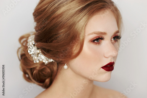 Beautiful young woman with blonde hair decorated with luxurious wedding accessory, pearl earrings and evening makeup. Portrait of attractive female model or bride with elegant hairstyle and red lips.