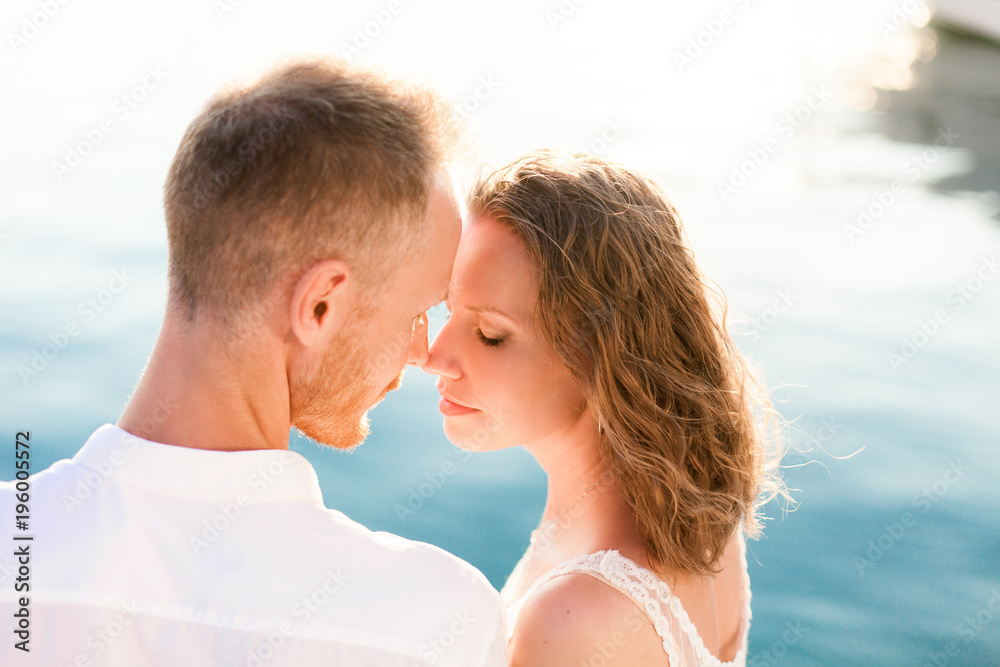 Couple of travelers are sitting by blue sea during romantic honeymoon vacation at sunset after wedding. Man and woman are happy tourists in resort. Lovers are wearing in white clothes outdoor. Couple of travelers are sitting by blue sea during romantic honeymoon vacation at sunset after wedding. Man and woman are happy tourists in resort. Lovers are wearing in white clothes outdoor.