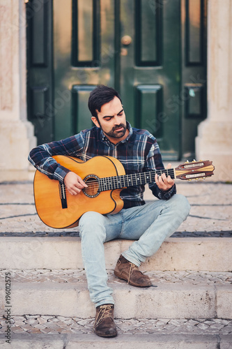 Stylish man with a beard in a plaid shirt and jeans in the street on the steps near the door. Macho with a beautiful smile sits in the evening on the bench and plays the guitar. Modern style of life.