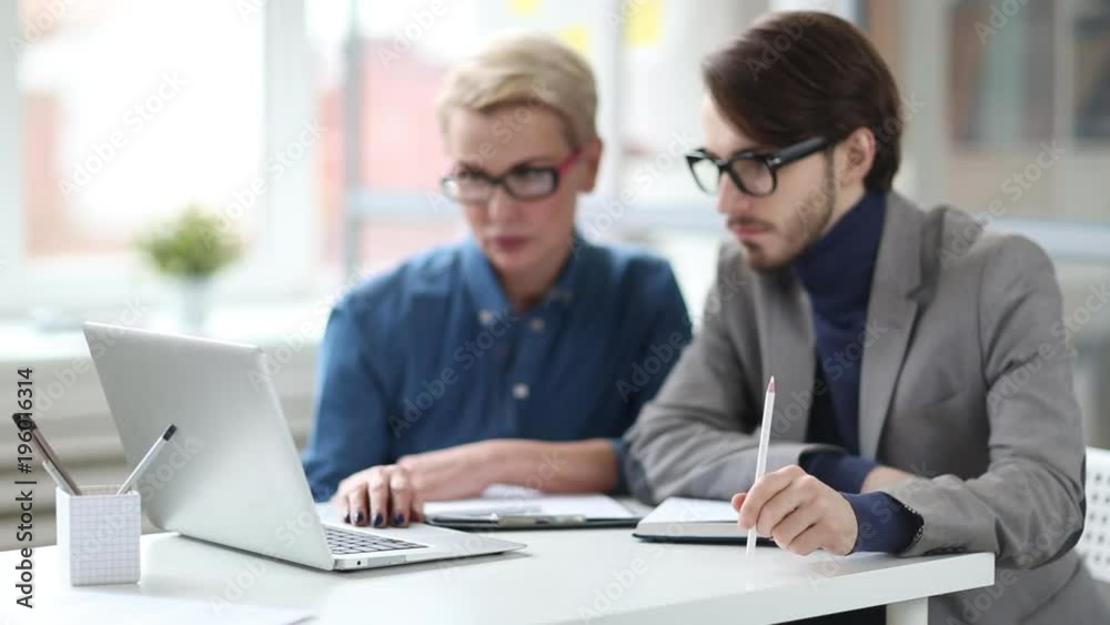 Group of hard-working managers gathered together in front of laptop and working on start-up project, bearded man tapping on table with pencil, office interior on background Group of hard-working managers gathered together in front of laptop and working on start-up project, bearded man tapping on table with pencil, office interior on background