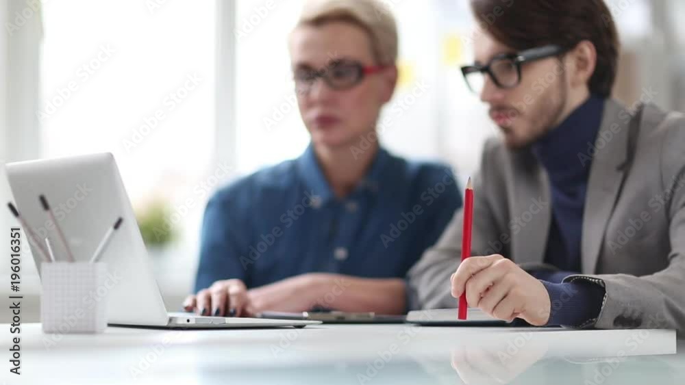 Young manager with stylish haircut sitting at office desk and showing results of accomplished work to handsome superior, he looking at laptop screen and tapping on table with pencil Young manager with stylish haircut sitting at office desk and showing results of accomplished work to handsome superior, he looking at laptop screen and tapping on table with pencil