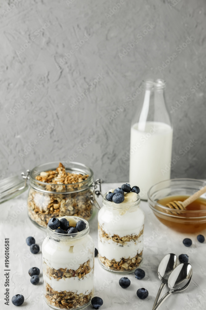 Homemade granola in a glass jar with yogurt and blueberries on a gray concrete background with a bottle of milk and honey. Food photography of a healthy morning breakfast.