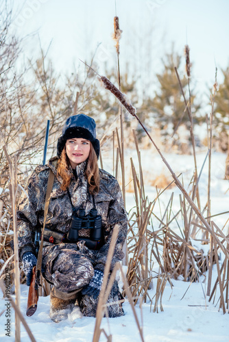 Beautiful girl hunter in camouflage suit with binoculars and a gun sits in the thickets of reeds, looking out for prey