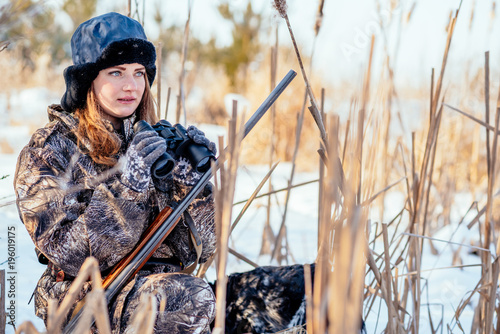 Beautiful girl hunter in camouflage suit with binoculars and a gun sits in the thickets of reeds, looking out for prey
