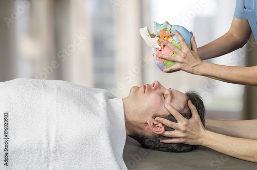 Physiotherapist doing a cranial sacral therapy to a man patient and another physiotherapist with a fake skull showing  position of the hands