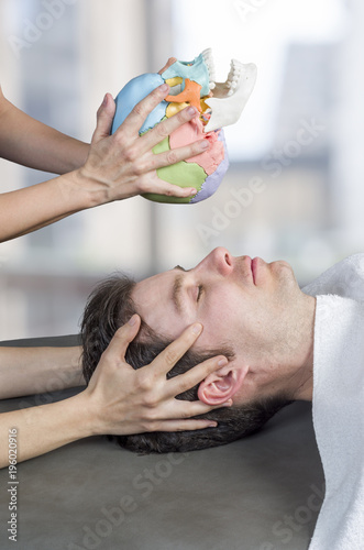 Physiotherapist doing a cranial sacral therapy to a man patient and another physiotherapist with a fake skull showing  position of the hands
