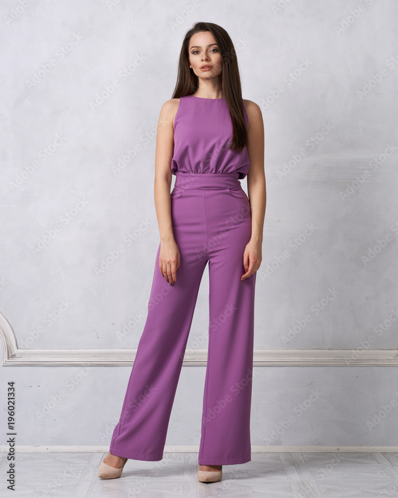 Agurk Far forår Charming female model with long brunette hair wearing fashionable  sleeveless purple jumpsuit and heeled shoes posing against white wall on  background. Gorgeous woman dressed in trendy apparel. Stock Photo | Adobe  Stock
