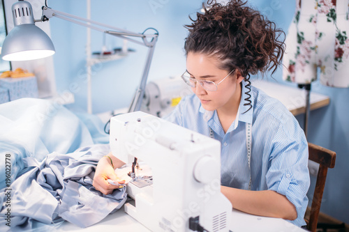 nice tailor with curly dark hair compliting work with sewing machine at workplace. neatly job. crative work. photo