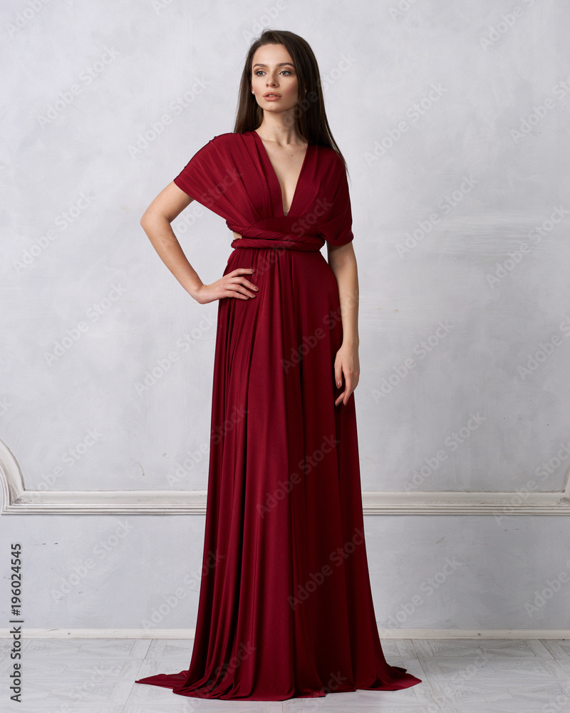 Beautiful long haired young woman dressed in stylish cherry red bandeau maxi dress posing against white wall on background. Elegant brunette female model demonstrating evening outfit in studio. Beautiful long haired young woman dressed in stylish cherry red bandeau maxi dress posing against white wall on background. Elegant brunette female model demonstrating evening outfit in studio.