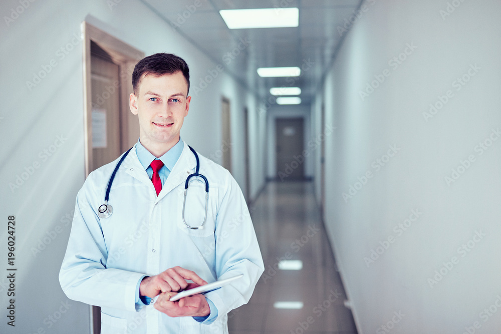 doctor in white medical coat is using a tablet and smiling while standing against window doctor in white medical coat is using a tablet and smiling while standing against window