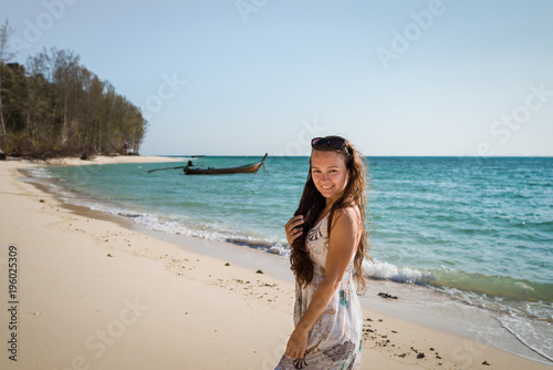 Young woman in dress is walking on the beach on the coast of the ocean. Smiling girl is enjoying sunny summer day and looking in camera. Tropical tavel destination