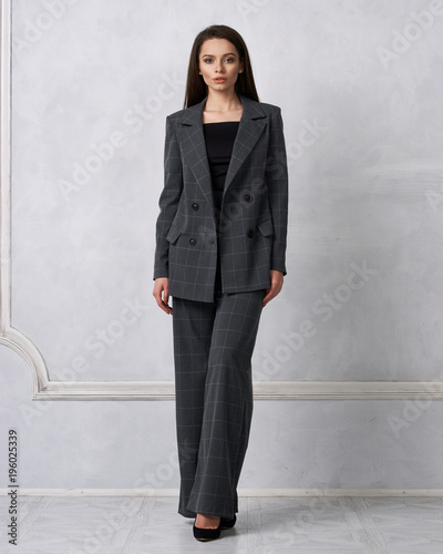 Gorgeous young woman dressed in gray squared jumpsuit, blazer and heeled shoes posing in studio. Beautiful brunette girl demonstrating stylish smart clothing against white wall on background. photo