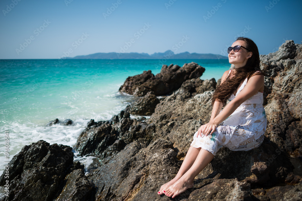 Young woman in dress sits on the rock on the coast of the ocean. Smiling girl is enjoying sunny summer day. Tropical tavel destination