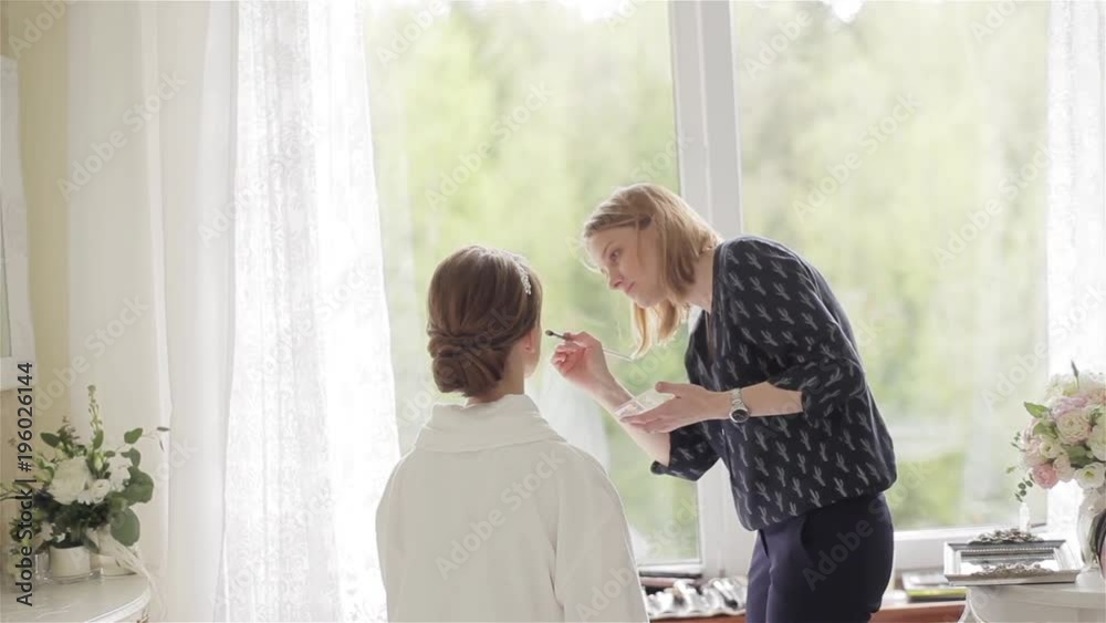 Professional make-up artist apply concealer using brush to face of model sitting back to camera by window at home or beauty salon natural lighting. No face woman getting ready prepared for photo shoot Professional make-up artist apply concealer using brush to face of model sitting back to camera by window at home or beauty salon natural lighting. No face woman getting ready prepared for photo shoot