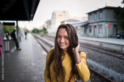 Young cute woman in bright dress stands on the train station and waits a train. Attractive brunette girl is smiling and looking in camera. Travel and adventures in Europe