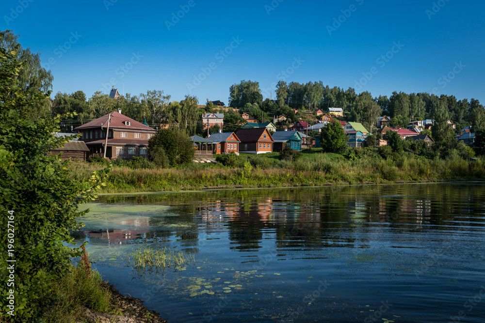 Scenic landscape with traditional buildings in Russian village on the riverbank. Background of nature with green trees, grass and hills. Clear sky