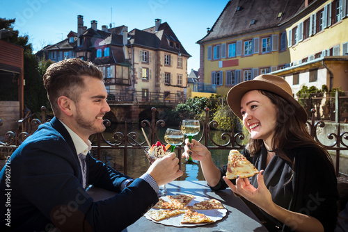 Young woman with boyfriend has lunch in Alsace. Two people sits together in local cafe with pizza and wine at the city promenade. Trendy couple holds wine glasses in hands on terrace in romantic place